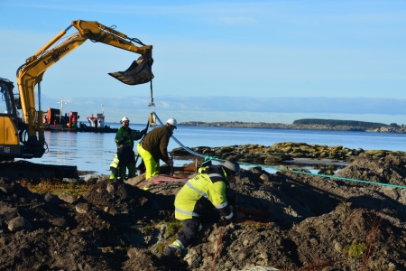 Montører legger ny kabel ved sjøen med hjelp av gravemaskin